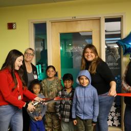 Adults and children in front of a closed door with red ribbon and scissors to conduct ribbon cutting and opening of the new sensory room.