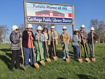 Photo of Library director & board members wearing hard hats and with gold shovels. Sign "Future home of the Carthage Public Library District"