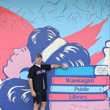 Artist stands in front of mural of a girl relaxing on the beach reading. He poses as if he is leaning on a stack of books where she rests her head.